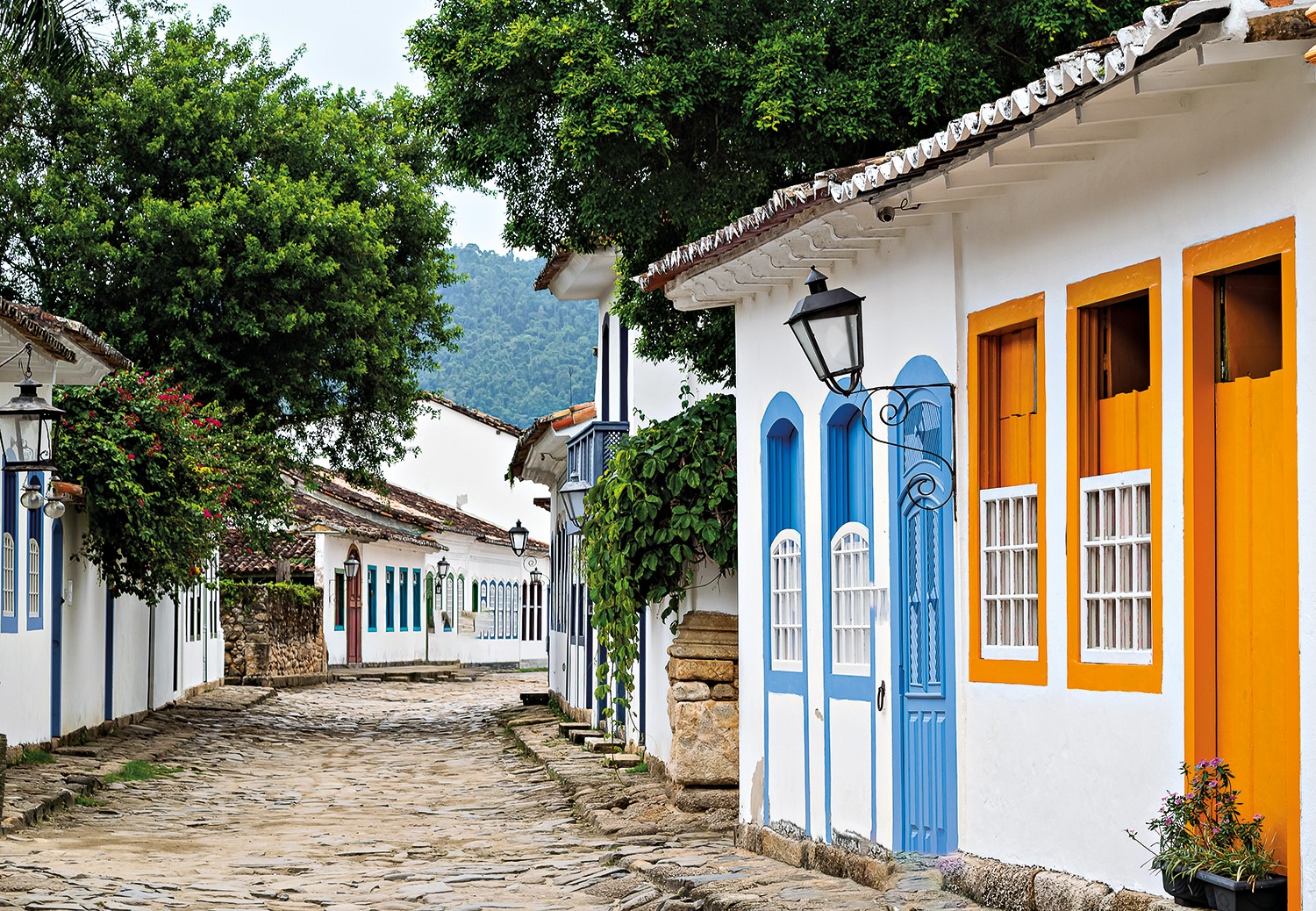 Rua de paralelep&iacute;pedos em Paraty, com casas coloniais brancas e telhados de barro. As janelas e portas s&atilde;o azuis e laranjas, e h&aacute; &aacute;rvores verdes ao fundo, com montanhas distantes