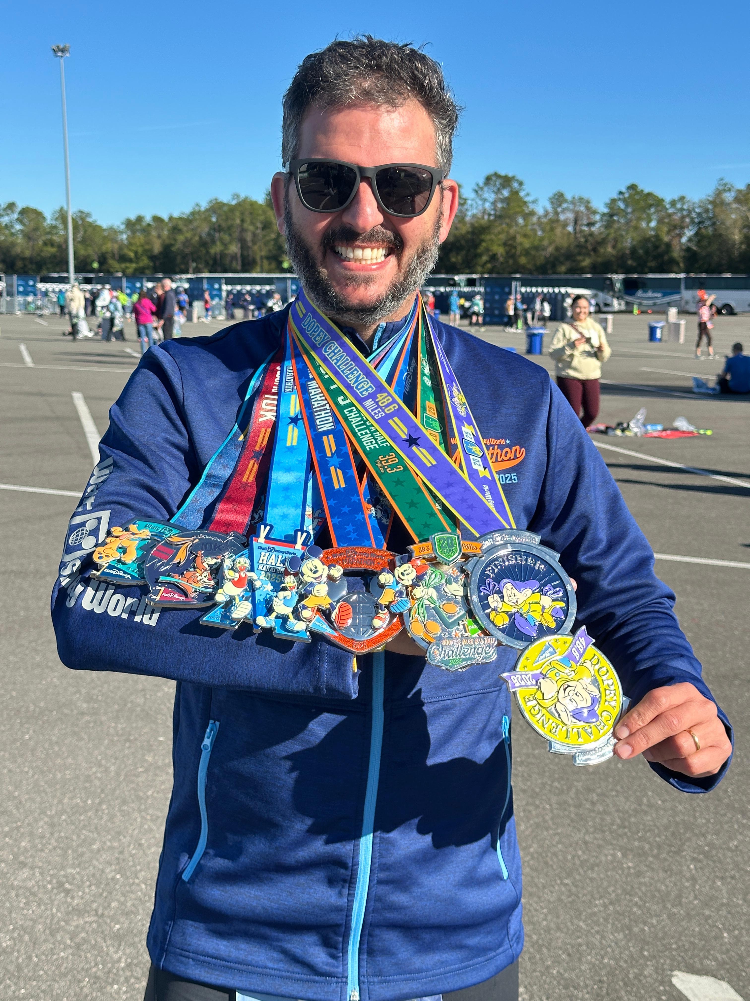 Homem branco de barba, &oacute;culos escuros e jaqueta azul sorri para a c&acirc;mera, exibindo v&aacute;rias medalhas de corrida com fitas coloridas e personagens da Disney. Ele est&aacute; em um estacionamento aberto, com outras pessoas e ve&iacute;culos ao fundo, sob um c&eacute;u azul claro