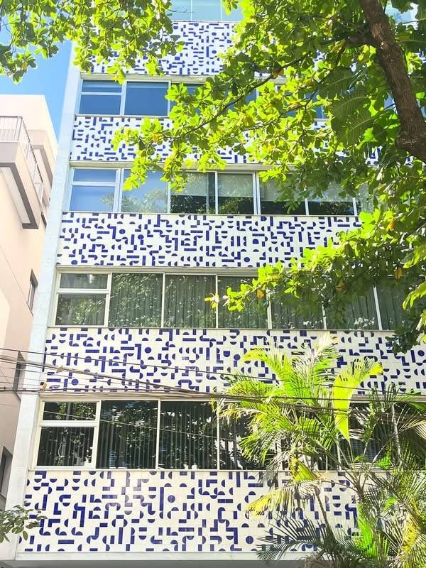 Edif&iacute;cio de v&aacute;rios andares com fachada branca e detalhes geom&eacute;tricos azuis, janelas amplas e &aacute;rvores frondosas em primeiro plano, sob c&eacute;u azul