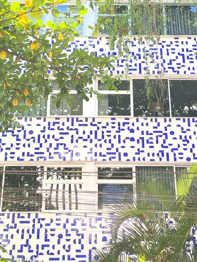 Fachada de edif&iacute;cio com azulejos brancos e padr&otilde;es geom&eacute;tricos azuis, como pontos e linhas. Janelas retangulares escuras refletem a vegeta&ccedil;&atilde;o, com folhas verdes e amareladas de &aacute;rvores em primeiro plano. Fios el&eacute;tricos cruzam a imagem.