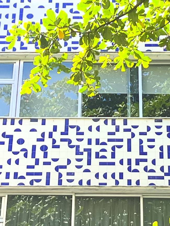 Fachada de edif&iacute;cio com azulejos brancos e padr&otilde;es geom&eacute;tricos azuis, como ret&acirc;ngulos e semic&iacute;rculos. Acima, janelas refletem &aacute;rvores e c&eacute;u. Galhos com folhas verdes vibrantes pendem sobre a parte superior da imagem, iluminados pelo sol