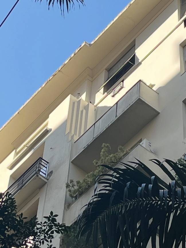 Fachada de um edif&iacute;cio bege de estilo Art D&eacute;co, com sacadas e janelas retangulares, sob c&eacute;u azul claro. Folhagens escuras de palmeiras em primeiro plano, &agrave; direita, e outras plantas verdes nas sacadas inferiores. Detalhes verticais em relevo na parede central.