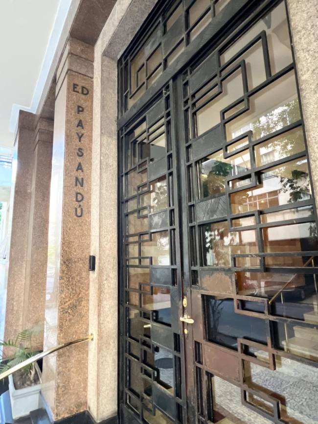 Entrada de um edif&iacute;cio com porta de vidro e grades pretas de design geom&eacute;trico. &Agrave; esquerda, uma coluna de granito marrom claro com o nome ED PAYSAND&Uacute; gravado verticalmente. Um corrim&atilde;o dourado e um vaso de planta verde complementam a cena