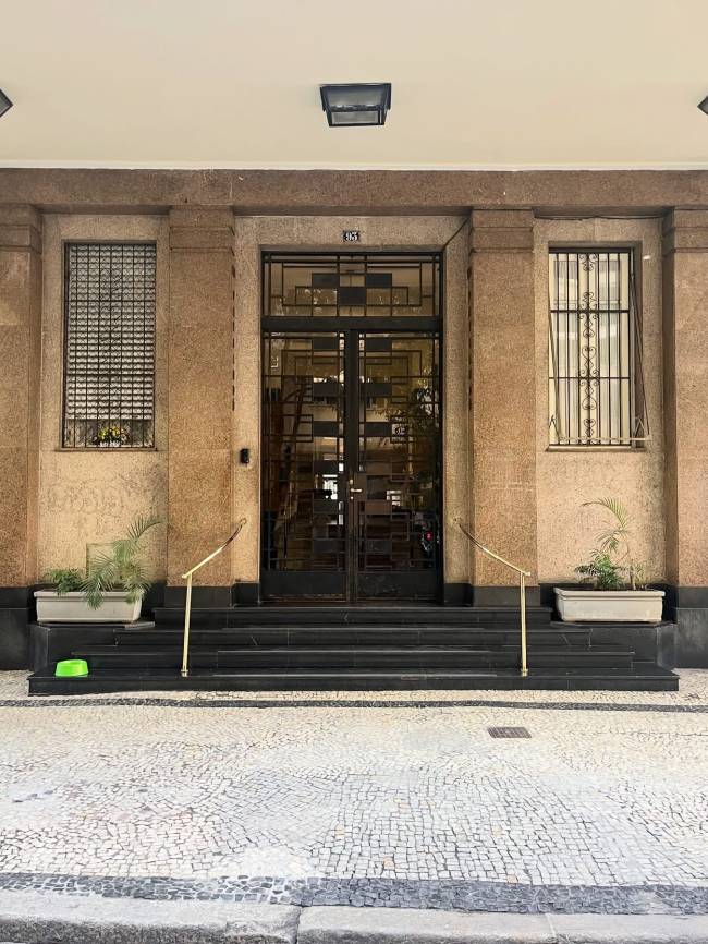 Fachada de edif&iacute;cio com porta dupla de ferro forjado, n&uacute;mero 135 acima, duas janelas laterais com grades, e tr&ecirc;s degraus pretos com corrim&atilde;os dourados. Vasos de plantas e um pote verde na cal&ccedil;ada de mosaico portugu&ecirc;s.