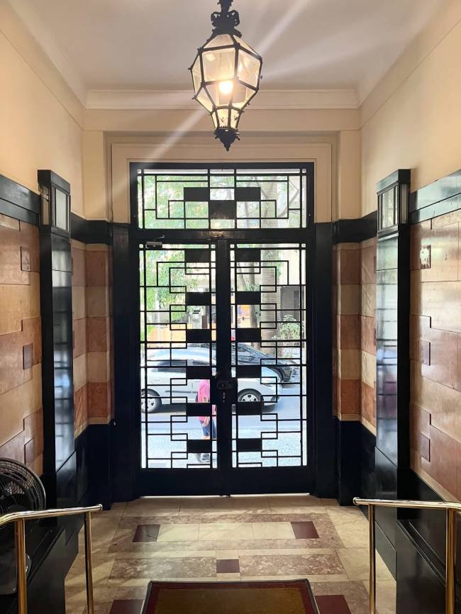 Corredor interno com piso de m&aacute;rmore claro e detalhes em marrom, paredes com faixas de azulejos bege e pretos, e um lustre de ferro forjado no teto. Ao fundo, uma porta de vidro com grades geom&eacute;tricas pretas revela a rua com carros e pessoas.