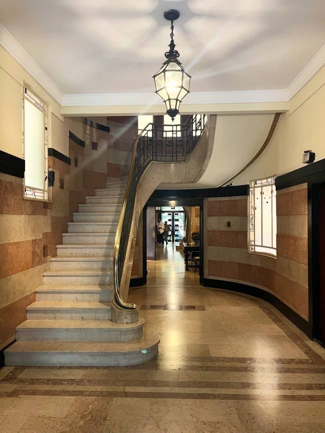 Hall de entrada com escadaria de m&aacute;rmore claro e corrim&atilde;o dourado &agrave; esquerda, levando a um andar superior com guarda-corpo de ferro. O piso &eacute; de m&aacute;rmore com detalhes em faixas escuras. As paredes t&ecirc;m revestimento em faixas horizontais de tons terrosos. Um lustre antigo de metal e vidro ilumina o centro do teto. Ao fundo, um corredor com pessoas e mesas.