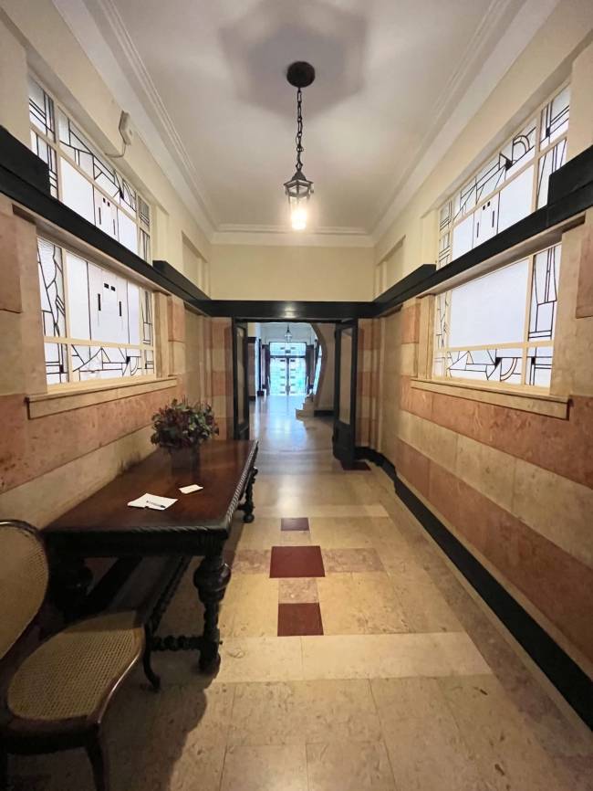 Corredor longo e iluminado com piso de m&aacute;rmore claro e detalhes em vermelho. As paredes t&ecirc;m faixas horizontais de m&aacute;rmore rosa e bege, com janelas superiores de vidro jateado e desenhos geom&eacute;tricos. Uma mesa de madeira escura com arranjo floral e uma cadeira de palha est&atilde;o &agrave; esquerda. Um lustre pendente ilumina o teto branco, e o corredor se estende at&eacute; uma porta aberta no fundo