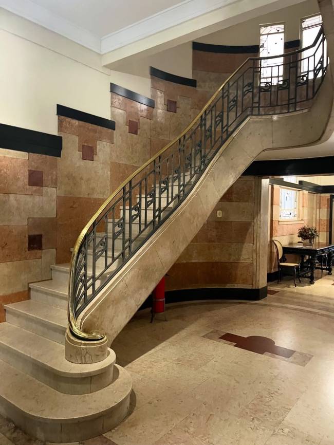 Escadaria elegante com degraus de m&aacute;rmore claro e corrim&atilde;o de ferro forjado com detalhes dourados. A parede &eacute; revestida com placas de m&aacute;rmore em tons de bege e marrom, intercaladas com faixas pretas. No canto direito, uma mesa escura com um vaso de flores e uma cadeira. O piso &eacute; de m&aacute;rmore claro com detalhes em marrom escuro