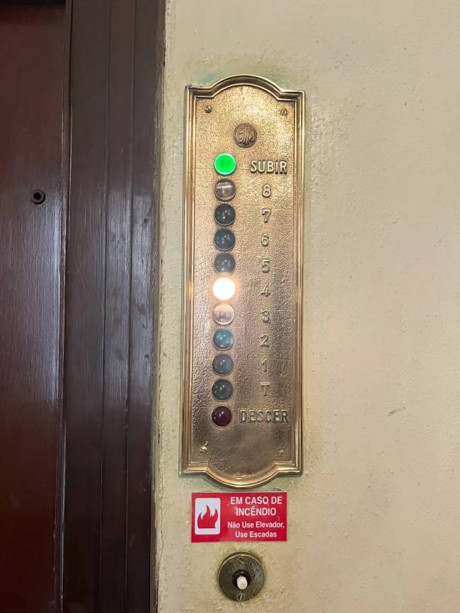 Painel de elevador antigo dourado com bot&otilde;es redondos para andares de 8 a T, e bot&otilde;es Subir e Descer. O bot&atilde;o Subir est&aacute; verde e o do andar 4 est&aacute; aceso em branco. Abaixo, um adesivo vermelho com um &iacute;cone de fogo e o texto EM CASO DE INC&Ecirc;NDIO N&atilde;o Use Elevador, Use Escadas