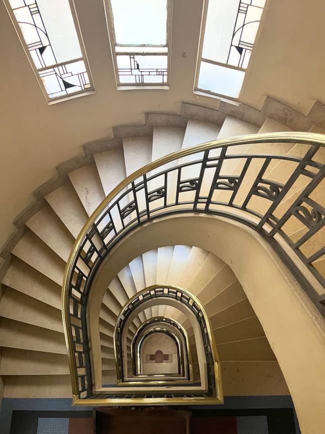 Vista de cima de uma escadaria em espiral com degraus de pedra clara e corrim&atilde;o de metal dourado e preto. No topo, tr&ecirc;s janelas retangulares com vitrais geom&eacute;tricos. A escada desce em c&iacute;rculos conc&ecirc;ntricos, revelando o piso do andar inferior com detalhes em azulejos azuis e marrons