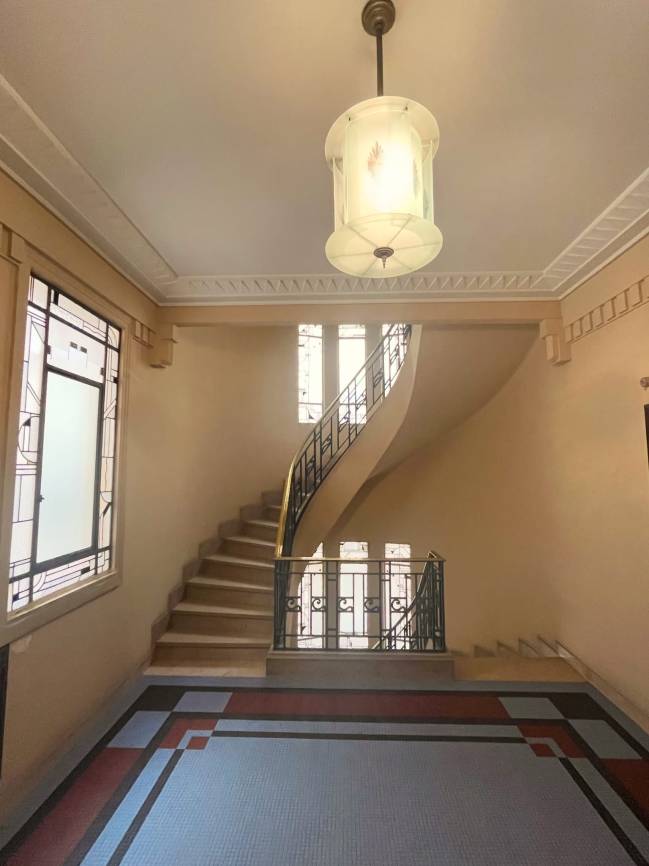 Hall de entrada com escada curva de degraus claros e corrim&atilde;o de ferro forjado preto com detalhes dourados. O piso tem mosaicos azuis, vermelhos e cinzas em padr&atilde;o geom&eacute;trico. Uma lumin&aacute;ria pendente de vidro fosco ilumina o teto com molduras decorativas. Janelas com vitrais geom&eacute;tricos filtram a luz natural