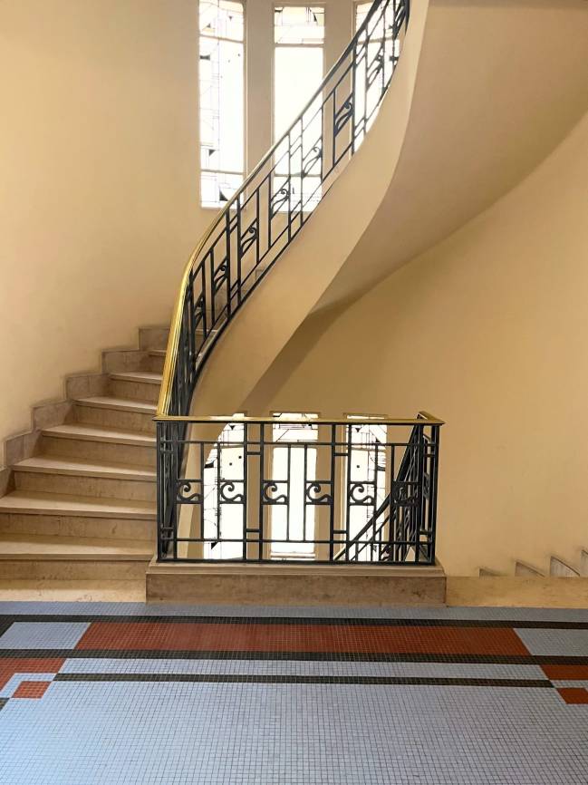 Escadaria curva de m&aacute;rmore bege com corrim&atilde;o de ferro forjado preto e dourado, paredes claras e piso de mosaico azul, preto e terracota. Janelas verticais no topo iluminam o ambiente