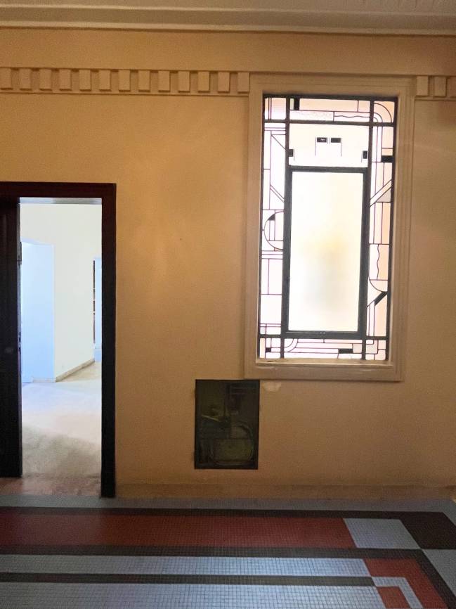 Corredor com paredes bege, porta escura aberta &agrave; esquerda revelando outro c&ocirc;modo claro, e janela alta com vitral geom&eacute;trico &agrave; direita. No ch&atilde;o, um mosaico de pequenos azulejos em tons de azul, vermelho e cinza forma um padr&atilde;o de faixas e &acirc;ngulos. Abaixo da janela, uma pequena caixa escura embutida na parede