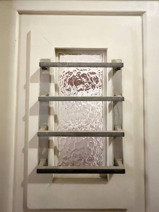 Detalhe de uma porta branca com um painel central retangular. No centro do painel, h&aacute; uma janela vertical com vidro texturizado, protegida por tr&ecirc;s barras horizontais de metal cinza escuro, espa&ccedil;adas uniformemente