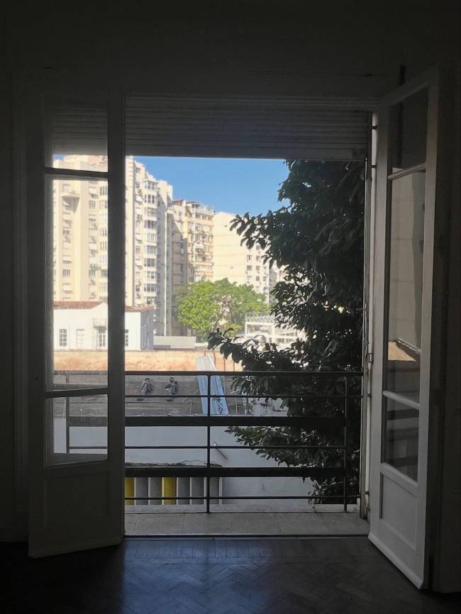 Vista de uma varanda com porta aberta para um cen&aacute;rio urbano. Edif&iacute;cios altos e uma &aacute;rvore frondosa preenchem o horizonte sob um c&eacute;u azul. Dois homens trabalham em um telhado pr&oacute;ximo. O ch&atilde;o da varanda &eacute; de madeira escura.