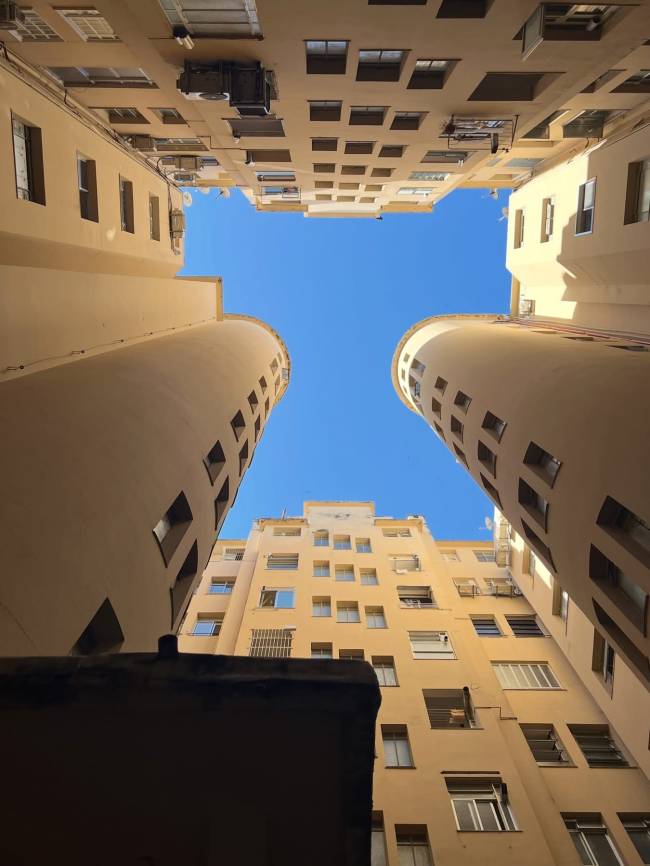 Vista de baixo para cima de um p&aacute;tio interno cercado por edif&iacute;cios altos de cor creme, com janelas retangulares e algumas redondas. O c&eacute;u azul claro &eacute; vis&iacute;vel no centro, emoldurado pelas paredes dos pr&eacute;dios que se elevam