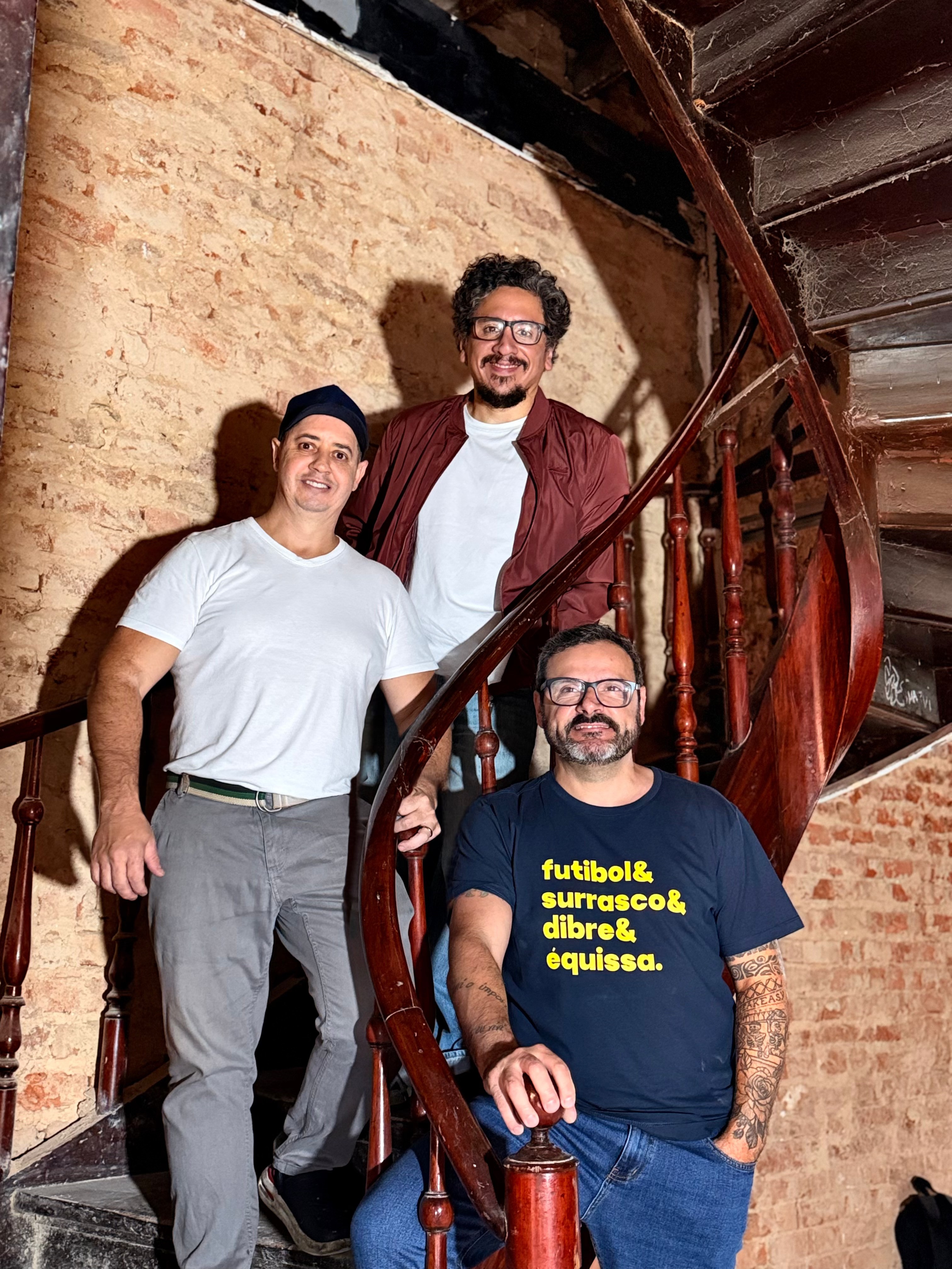 Três homens sorrindo em uma escada caracol de madeira escura. Um usa camiseta branca e gorro preto, outro camiseta branca e jaqueta vinho, e o terceiro camiseta azul com a frase futebol & churrasco & drible & equissa em amarelo