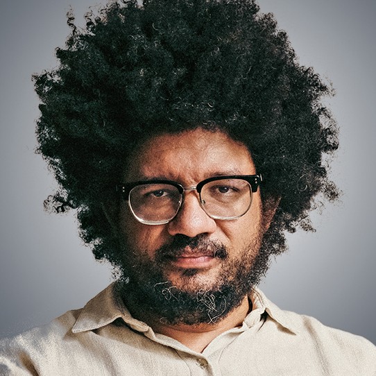 Homem negro de pele escura, cabelo crespo volumoso e barba cheia, usando &oacute;culos de arma&ccedil;&atilde;o preta e camisa bege, olhando s&eacute;rio para frente