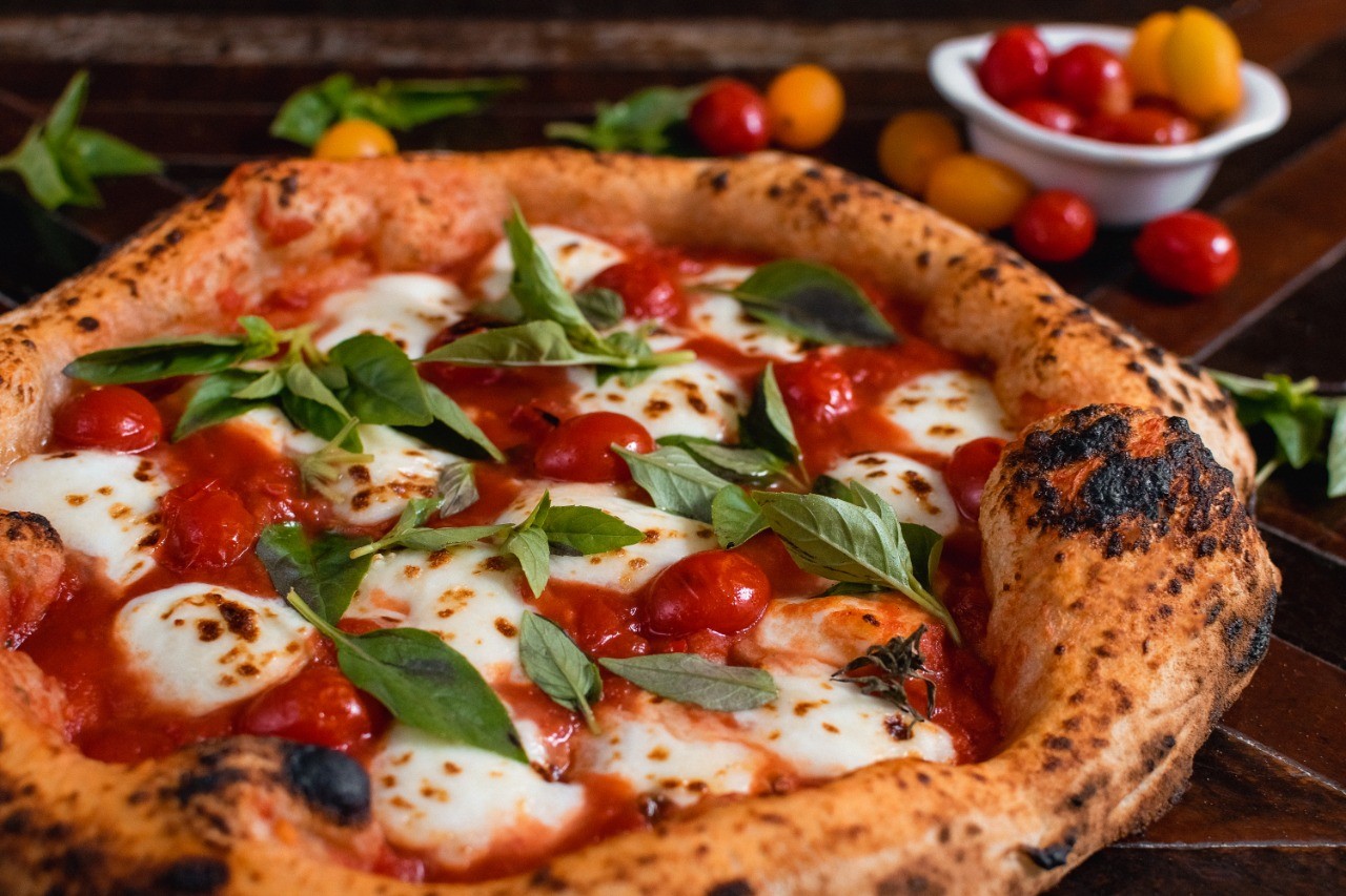 Pizza margherita com borda alta e crocante, molho de tomate, queijo mussarela em rodelas e folhas de manjeric&atilde;o fresco. Ao fundo, tomates cereja vermelhos e amarelos em uma tigela branca e espalhados sobre uma mesa de madeira escura