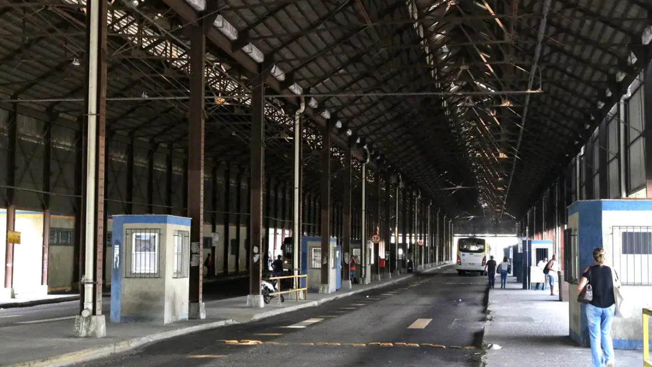 Interior de um terminal rodoviário antigo, com teto alto de estrutura metálica e telhas escuras. Um ônibus branco está parado ao fundo, e algumas pessoas aguardam ou caminham pela plataforma. Cabines de cor bege com detalhes azuis estão dispostas ao longo do corredor central, que possui faixas de pedestres no chão. A iluminação é natural, vinda das laterais e do fundo