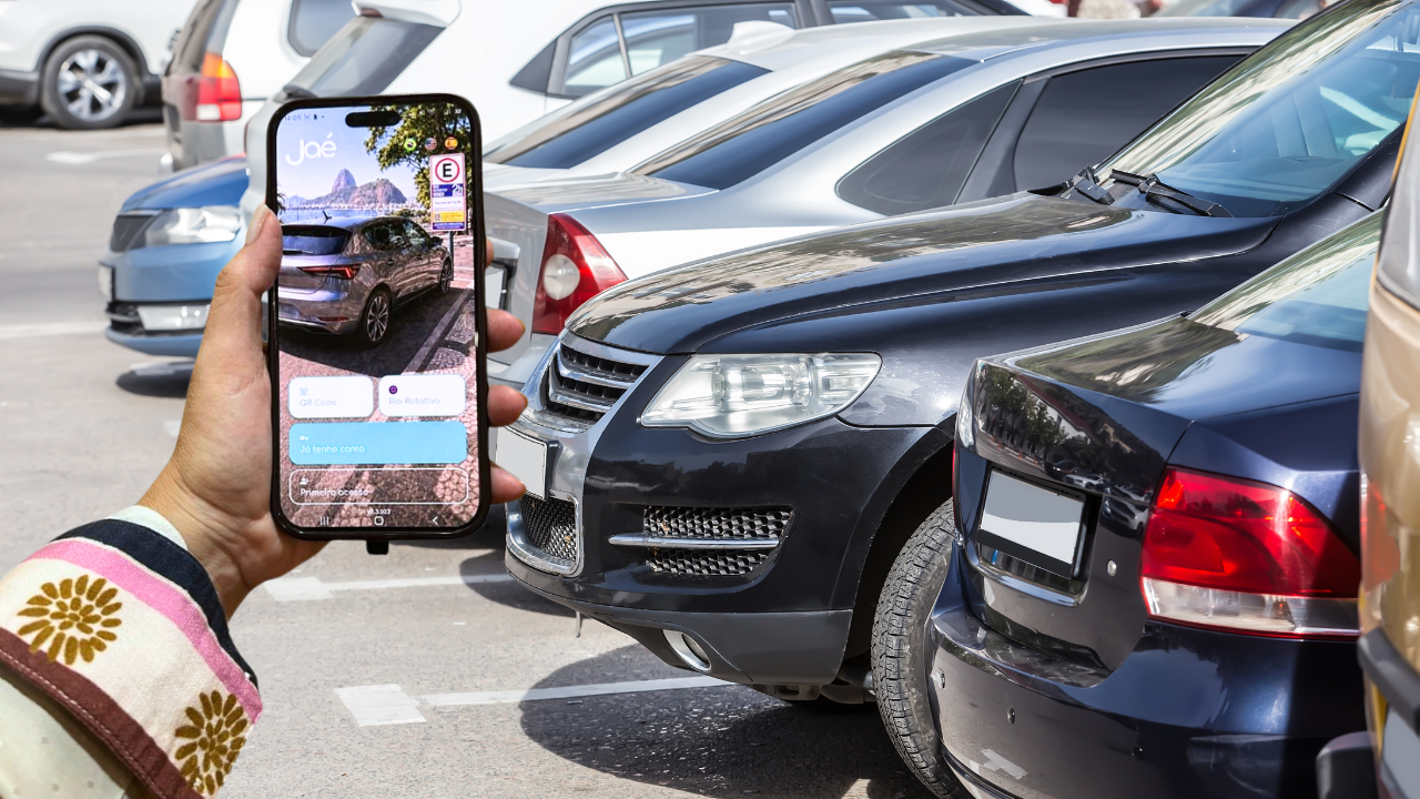 Mão segurando um smartphone com o aplicativo Joé aberto, mostrando um carro cinza estacionado e o Pão de Açúcar ao fundo. O celular está sobre um estacionamento com vários carros, incluindo um preto e um azul escuro