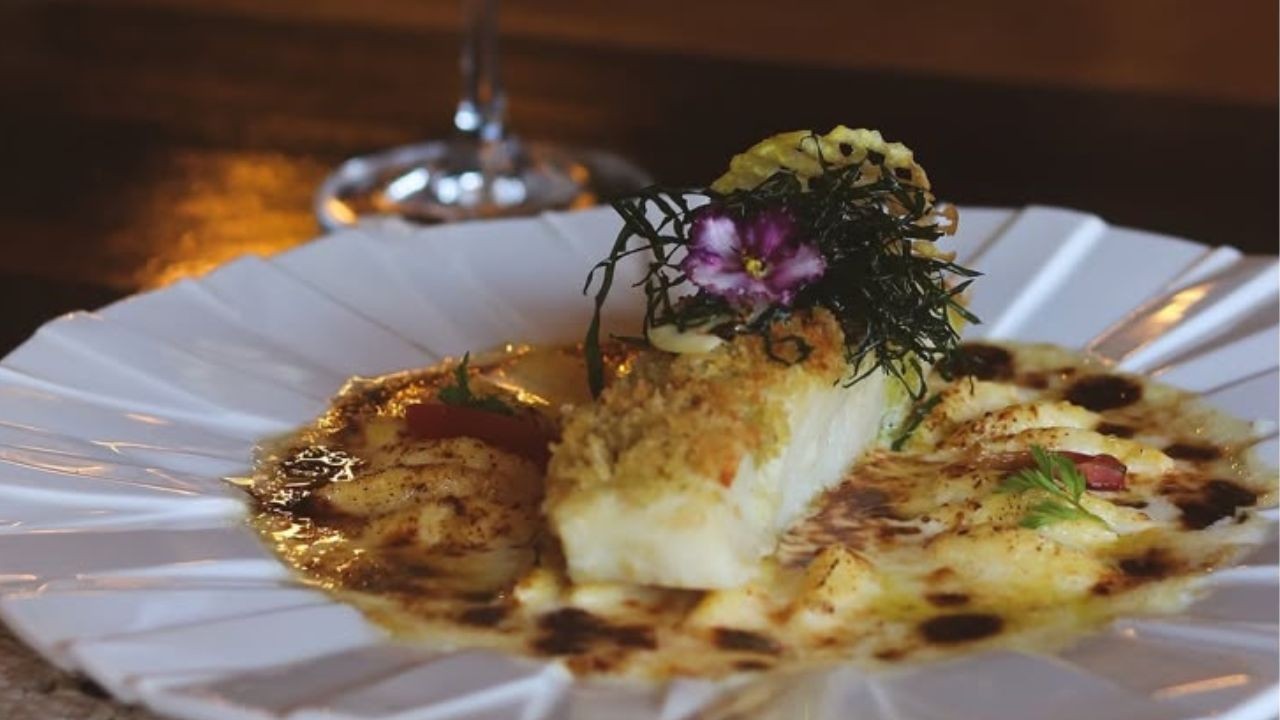 Prato gourmet com um peda&ccedil;o de peixe branco empanado, coberto por algas escuras e uma flor roxa, servido sobre um molho cremoso com pontos escuros e um peda&ccedil;o de piment&atilde;o vermelho, em um prato branco com bordas onduladas. Ao fundo, um copo de vinho desfocado
