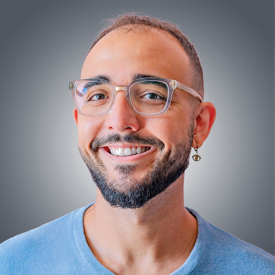 Homem branco de barba e &oacute;culos claros, sorrindo para a c&acirc;mera, com brinco de olho no l&oacute;bulo direito, vestindo camiseta azul