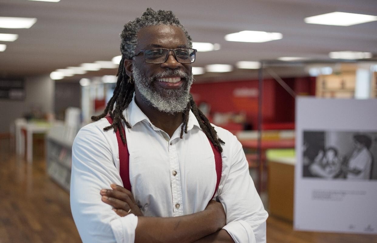 Homem negro de dreadlocks grisalhos e barba branca, sorrindo, usando &oacute;culos, camisa branca e suspens&oacute;rios vermelhos, bra&ccedil;os cruzados, em ambiente interno com ilumina&ccedil;&atilde;o no teto e exposi&ccedil;&otilde;es ao fundo