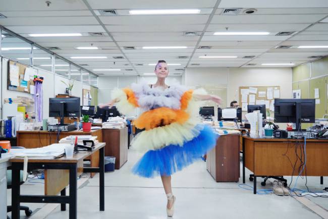 Mulher sorridente, com maquiagem colorida no rosto e cabelo curto, dan&ccedil;a bal&eacute; em ponta no centro de um escrit&oacute;rio. Ela veste um tutu volumoso de tule em camadas brancas, laranjas e azuis, com um top preto. Mesas de madeira com monitores e pap&eacute;is desorganizados preenchem o fundo. Um homem trabalha em uma das mesas &agrave; direita