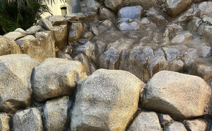 Uma cascata artificial de pedras grandes e m&eacute;dias em tons de cinza e marrom, com &aacute;gua escorrendo sobre elas, criando um efeito molhado e brilhante. Ao fundo, folhagens verdes e uma constru&ccedil;&atilde;o clara aparecem parcialmente