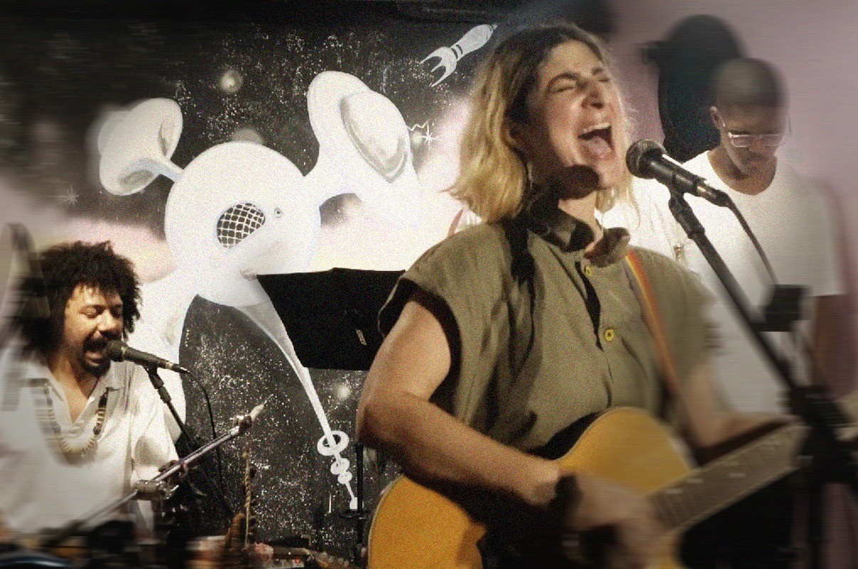 Cantora loira de camisa verde-oliva tocando viol&atilde;o e cantando com a boca aberta, ao lado de um homem de cabelo crespo e barba cantando em um microfone, com um fundo escuro e desenhos abstratos brancos