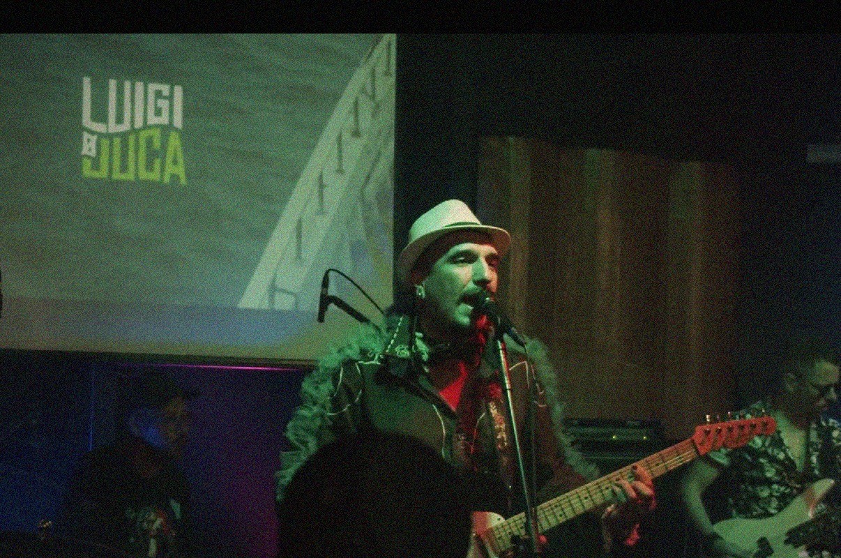 Cantor de chap&eacute;u branco e casaco peludo verde, com barba e bigode, canta no microfone e toca guitarra em palco escuro. Ao fundo, proje&ccedil;&atilde;o verde com o nome LUIGI E LUCA. Outros m&uacute;sicos aparecem desfocados.