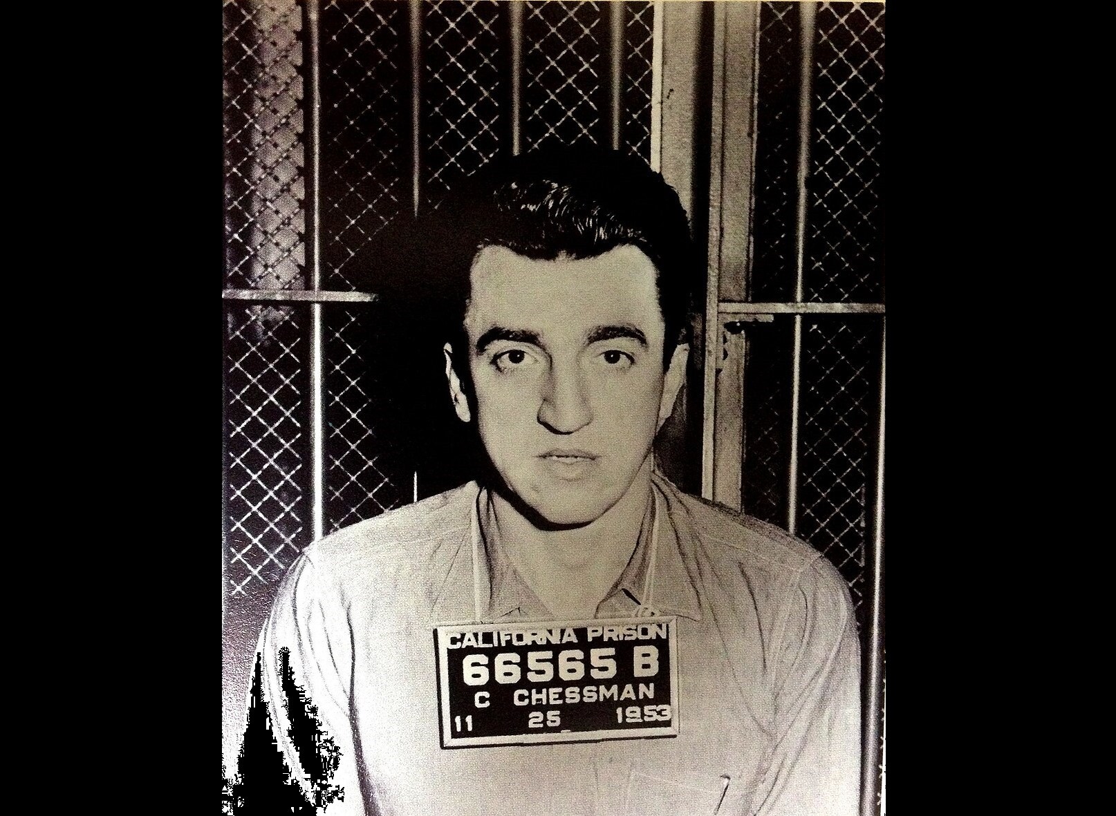 Homem branco de cabelo escuro, com expressão séria, usando camisa clara e uma placa de identificação de prisão com CALIFORNIA PRISON 66565 B C CHESSMAN 11 25 1953 no peito, em frente a grades