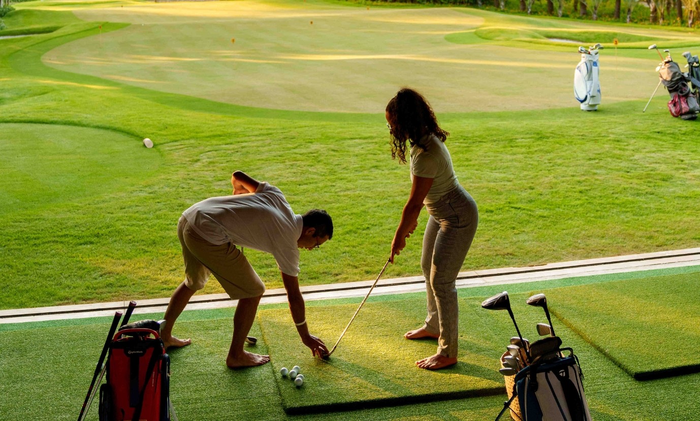 Um homem e uma mulher descalços praticam golfe em um campo verde. O homem, de camisa e bermuda claras, se curva para posicionar uma bola. A mulher, de camiseta e calça cinzas, segura um taco, observando. Bolsas de golfe estão ao lado, e o gramado se estende ao fundo