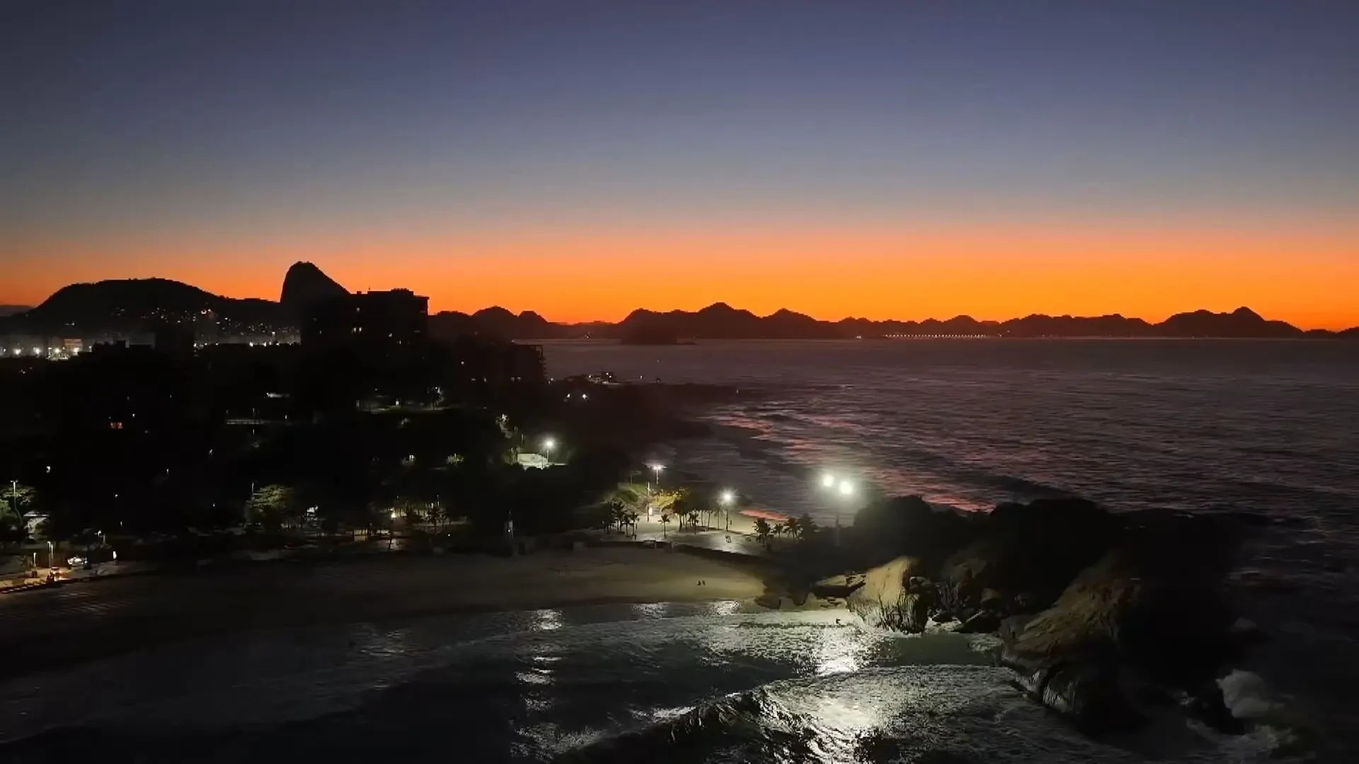 Vista aérea noturna da orla do Rio de Janeiro, com o Pão de Açúcar e outros morros ao fundo. O céu exibe um gradiente de azul escuro a laranja vibrante no horizonte. A cidade está escura, com luzes pontuais iluminando a praia e o calçadão. O mar reflete as luzes, e ondas quebram na areia e nas rochas. Montanhas escuras se estendem ao longo da costa distante