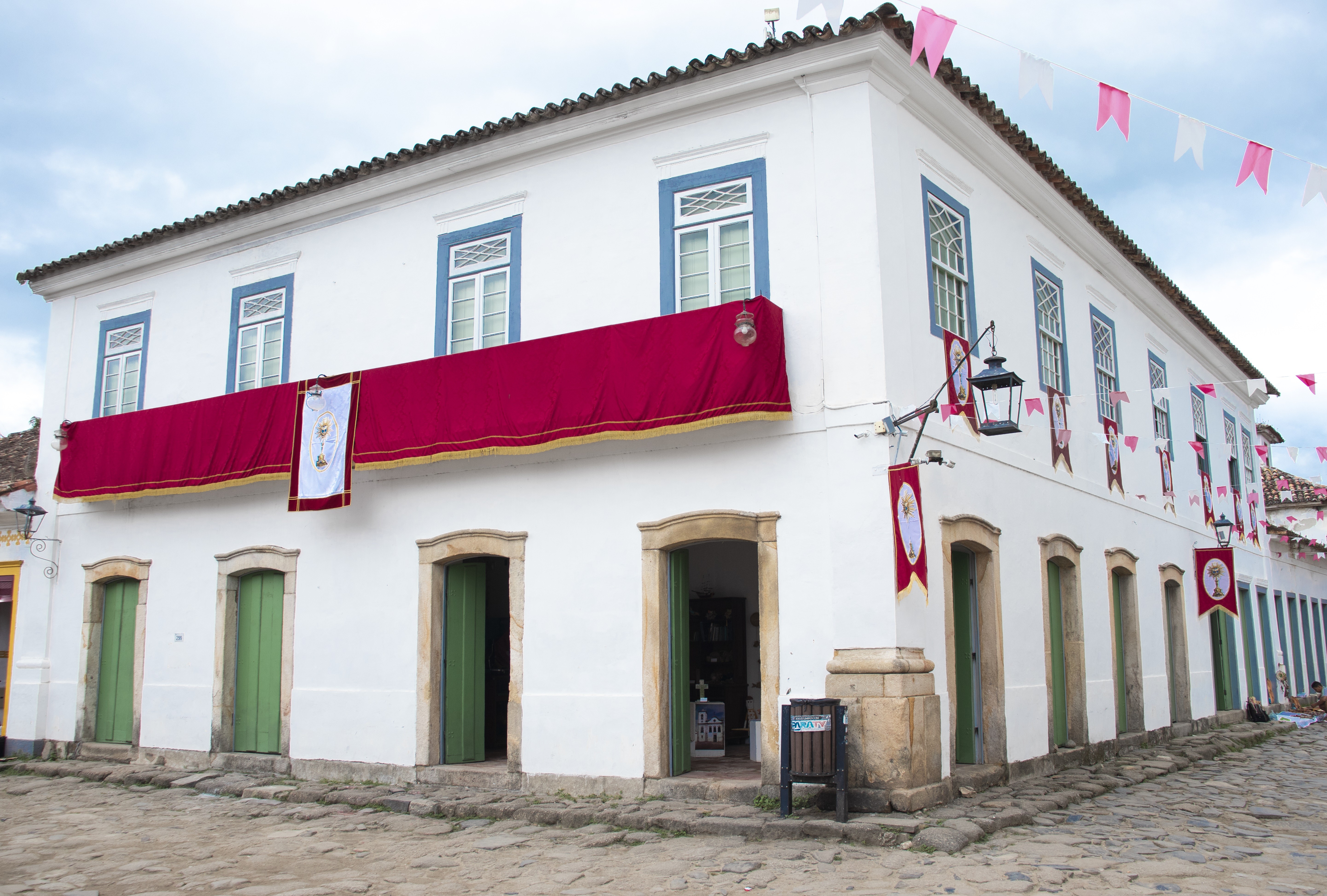 Casar&atilde;o colonial branco com janelas azuis e portas verdes, decorado com um tecido vermelho no andar superior e bandeirinhas rosa e brancas penduradas no c&eacute;u, em uma rua de paralelep&iacute;pedos