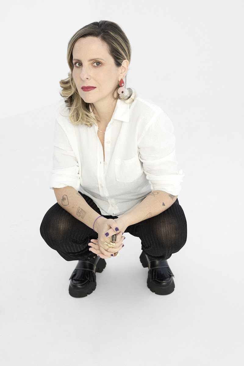 Mulher de cabelos loiros e castanhos, batom vermelho, camisa branca e cal&ccedil;a preta, agachada com as m&atilde;os unidas sobre os joelhos. Ela usa brincos grandes, an&eacute;is dourados e sapatos pretos de plataforma, exibindo tatuagens nos bra&ccedil;os