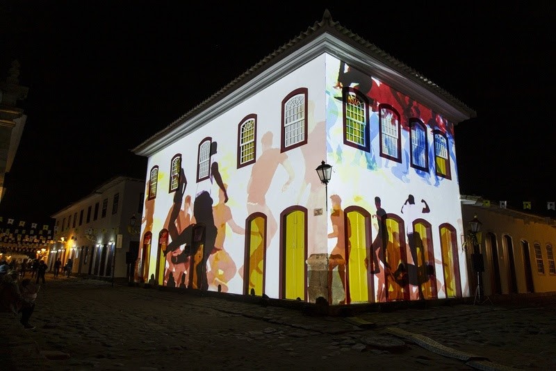 Fachada de um casar&atilde;o colonial branco iluminada por proje&ccedil;&otilde;es coloridas de silhuetas humanas dan&ccedil;ando, com janelas e portas emolduradas em amarelo. A rua de paralelep&iacute;pedos e outros pr&eacute;dios escuros ao fundo criam um ambiente noturno festivo
