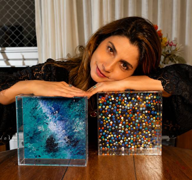 Mulher de cabelos castanhos e blusa preta de renda sorri, apoiando os bra&ccedil;os sobre duas caixas de acr&iacute;lico. A caixa &agrave; esquerda exibe uma pintura abstrata em tons de azul e verde, enquanto a da direita cont&eacute;m pequenas esferas coloridas.