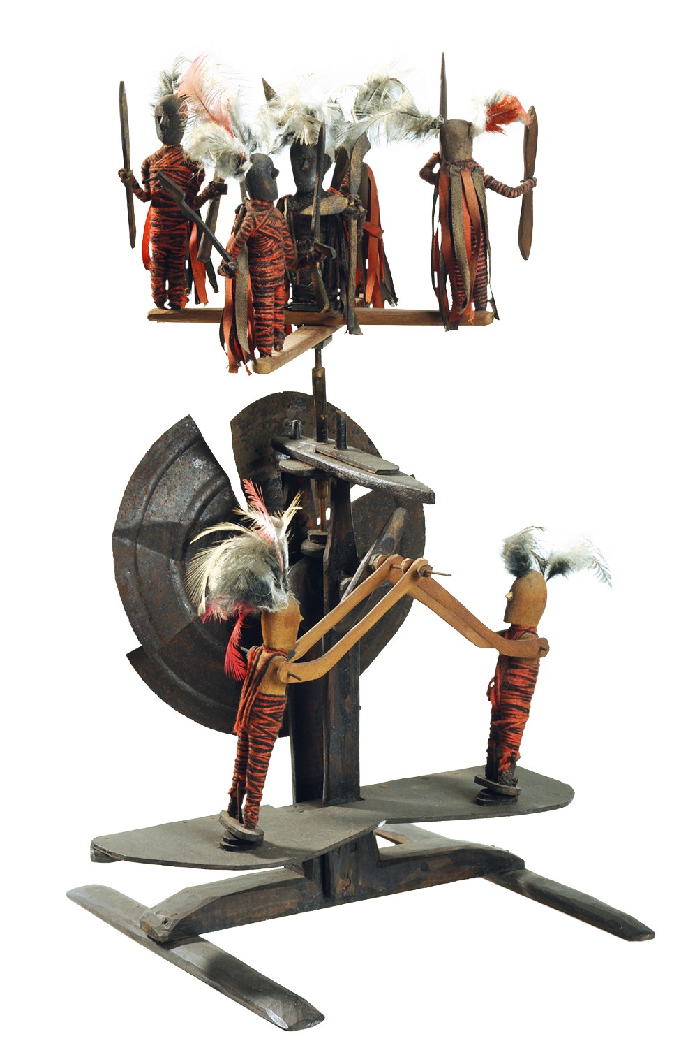 Escultura de madeira escura com figuras humanas estilizadas. Na parte superior, cinco figuras com corpos envoltos em tecido laranja e branco, adornadas com penas brancas e marrons, seguram objetos pontiagudos. Abaixo, duas figuras semelhantes, com penas e tecidos, operam uma estrutura de madeira que se conecta a um disco met&aacute;lico. A base &eacute; de madeira escura e plana