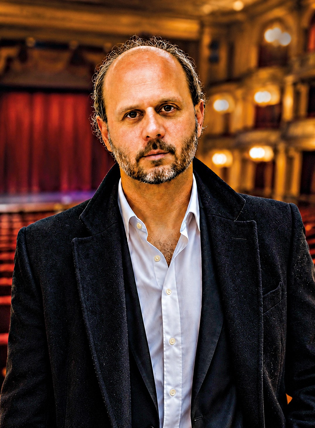 Homem branco de barba e cabelo ralo, vestindo casaco escuro e camisa branca aberta, em p&eacute; num teatro com poltronas vermelhas e palco ao fundo