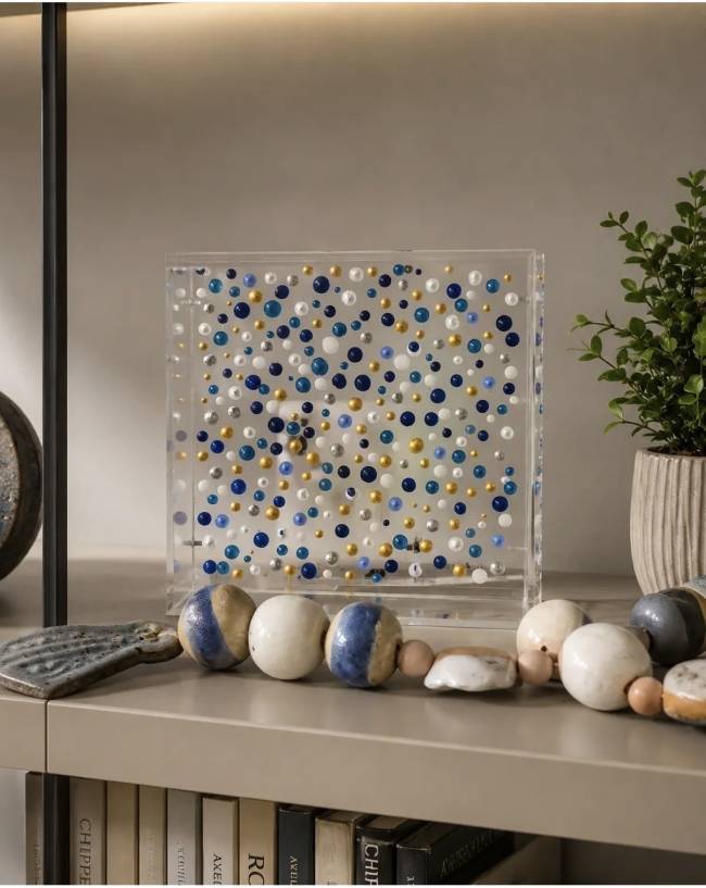 Decora&ccedil;&atilde;o de estante com caixa de acr&iacute;lico transparente cheia de bolinhas azuis, brancas e douradas, ao lado de um vaso com planta verde e um colar de contas de cer&acirc;mica em tons de azul, branco e bege. Livros com lombadas claras e escuras preenchem a prateleira inferior