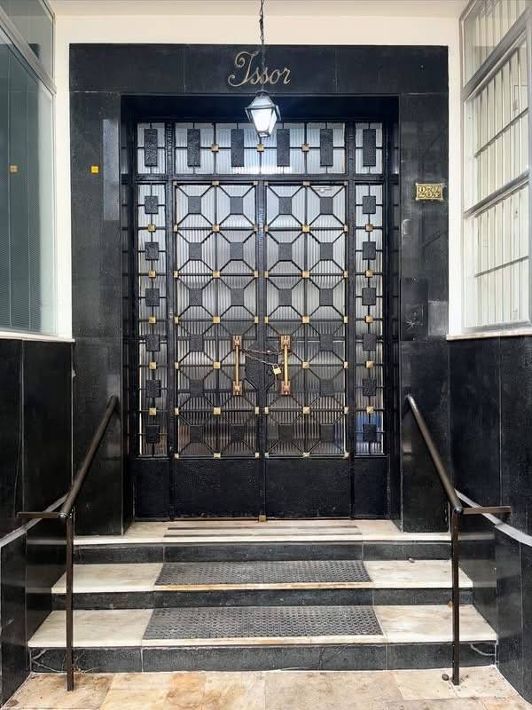 Entrada de pr&eacute;dio com porta dupla de ferro forjado preto e detalhes dourados, vidro canelado, e letreiro Tssor acima. Degraus de m&aacute;rmore preto e branco com corrim&atilde;os escuros, e um lustre pendurado iluminando a porta
