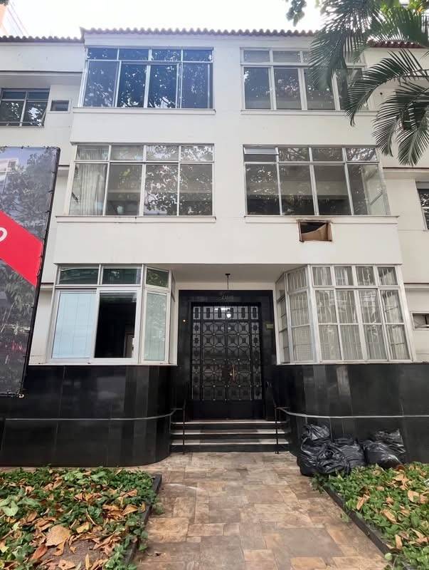 Fachada de um edif&iacute;cio residencial branco de tr&ecirc;s andares com janelas amplas e sacadas envidra&ccedil;adas. No t&eacute;rreo, uma porta preta ornamentada com detalhes geom&eacute;tricos, ladeada por janelas e revestimento escuro. Em frente, um caminho de pedras e canteiros com folhagens verdes e secas. Sacos de lixo pretos est&atilde;o &agrave; direita da entrada. Uma palmeira aparece no canto superior direito