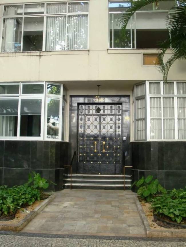 Entrada de um pr&eacute;dio com fachada bege, janelas amplas e uma porta dupla preta com detalhes dourados e gradeado geom&eacute;trico. H&aacute; tr&ecirc;s degraus e corrim&atilde;os de metal, ladeados por canteiros com folhagens verdes e um caminho de pedras claras. Uma palmeira aparece no canto superior direito