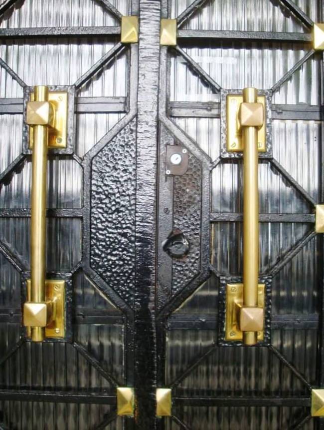 Port&atilde;o duplo de ferro forjado preto com detalhes dourados, incluindo duas ma&ccedil;anetas verticais e ornamentos quadrados. O centro tem uma fechadura e textura martelada, enquanto as laterais exibem um padr&atilde;o geom&eacute;trico sobre um fundo listrado verticalmente
