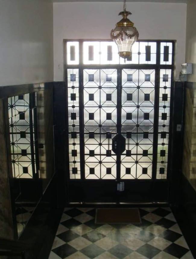 Porta de entrada escura com detalhes em vidro texturizado e grades geom&eacute;tricas, iluminada por luz externa. Um lustre antigo de bronze e vidro pendurado no teto. O ch&atilde;o tem um padr&atilde;o xadrez preto e branco, com um tapete marrom &agrave; frente da porta. Paredes laterais escuras refletem a porta, criando um efeito de espelho