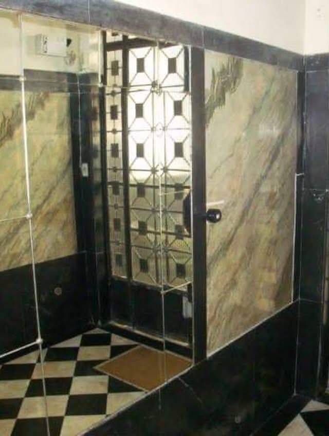 Corredor com piso xadrez preto e branco, paredes com espelhos e m&aacute;rmore bege, refletindo uma porta de ferro com detalhes geom&eacute;tricos e um tapete marrom no ch&atilde;o