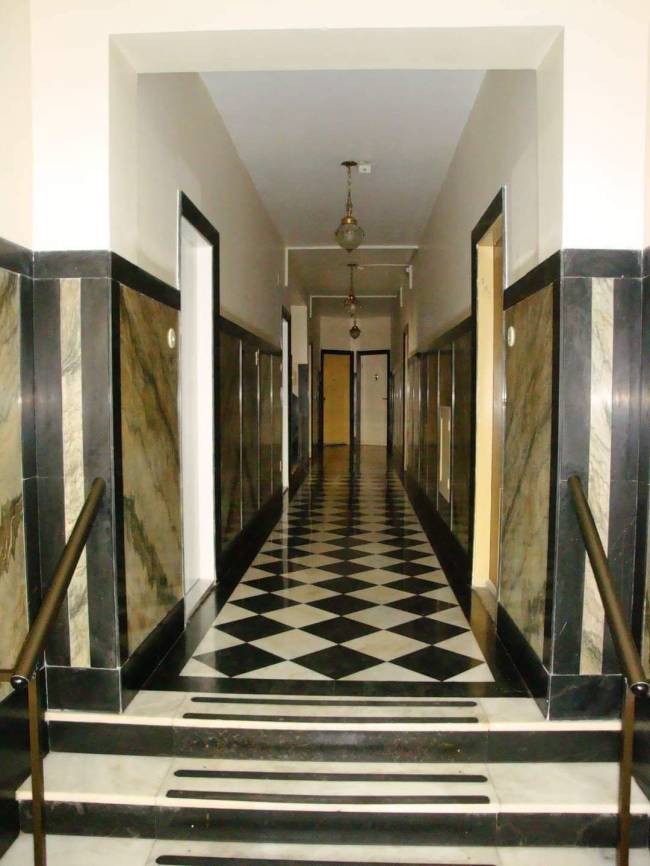 Corredor longo com piso xadrez preto e branco, paredes revestidas de m&aacute;rmore escuro e claro, e teto branco com lumin&aacute;rias pendentes. H&aacute; degraus de m&aacute;rmore preto e branco com faixas antiderrapantes e corrim&atilde;os escuros nas laterais, levando ao corredor com portas vis&iacute;veis ao fundo