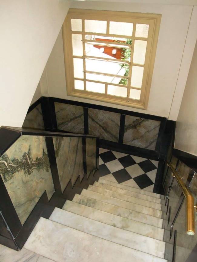 Escadaria de m&aacute;rmore claro com corrim&atilde;o dourado, paredes laterais revestidas em m&aacute;rmore verde e preto, e um patamar com piso xadrez preto e branco. Acima, uma janela retangular com grades brancas e vista para folhagem verde